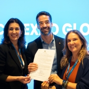 Uma foto de três pessoas (duas mulheres e um homem, no centro) em um evento, posando e segurando um documento (possivelmente um termo de cooperação ou parceria). Eles estão sorrindo. O homem está de terno escuro, e as mulheres vestem blazers pretos ou escuros sobre roupas em tons quentes (uma com gola branca, a outra com tom alaranjado). O fundo é um painel de LED ou projeção em tom azul-claro, com parte do texto em branco que diz "CONEXAO GLOBAL" e mais abaixo, parcialmente visível, "Tória d". A iluminação é de evento, com foco nas pessoas.