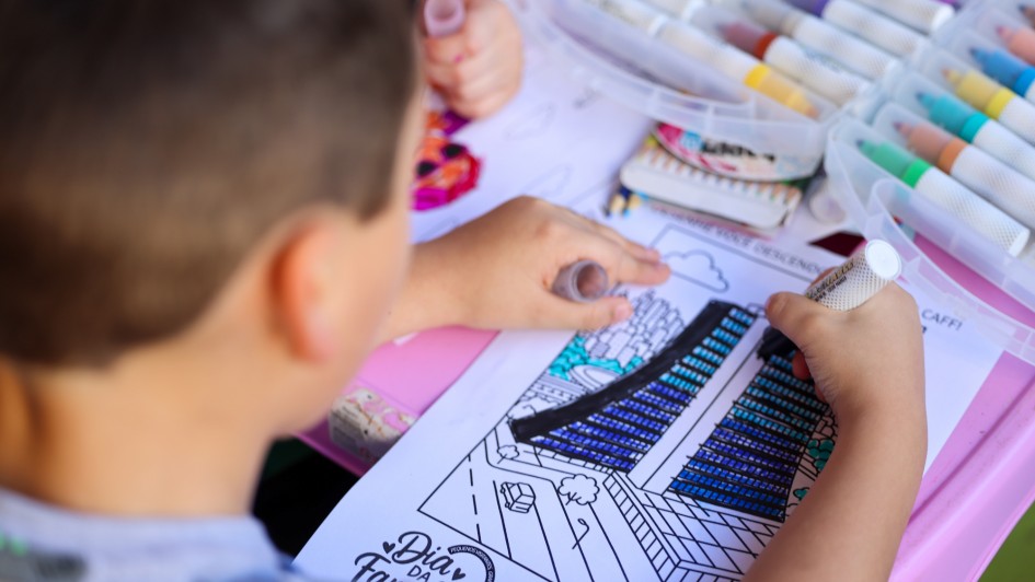Foto de uma criança, que aparece de costas, colorindo um desenho do Caff. A mesa usada é colorida e apresenta diversas canetinhas de colorir.