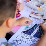 Foto de uma criança, que aparece de costas, colorindo um desenho do Caff. A mesa usada é colorida e apresenta diversas canetinhas de colorir.