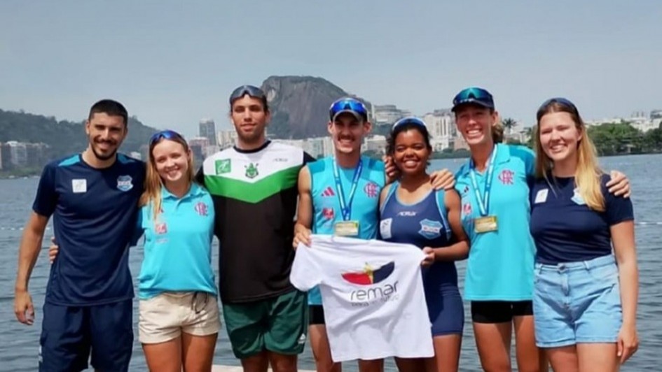 A imagem apresenta um grupo de pessoas em pé, posicionado à beira de um lago, com uma paisagem urbana ao fundo. Todos vestem roupas esportivas e exibem medalhas penduradas no pescoço. No centro, uma pessoa segura uma camiseta branca com o logotipo “Remar para o Futuro”.