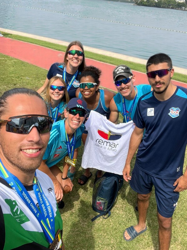 A foto mostra um grupo de pessoas reunidas em área externa gramada, próxima a um corpo d’água com pista de remo ao fundo. O grupo veste uniformes esportivos em tons de azul e segura uma camiseta branca com o logotipo “Remar para o Futuro” estampado no centro. Algumas pessoas usam medalhas penduradas no pescoço.