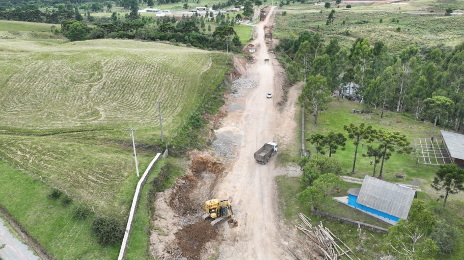 Uma foto aérea ou em ângulo elevado mostrando uma estrada rural de terra em construção ou manutenção (ERS-427). A estrada de cor clara, com sulcos e terraplanagem em andamento, corta a paisagem de campos verdes e árvores. No primeiro plano, à esquerda da estrada, há uma escavadeira amarela trabalhando, removendo terra e detritos. Um caminhão basculante está parado mais à frente na estrada. Ao longe, no meio da estrada, um veículo de cor clara está transitando. A área é predominantemente verde, com colinas suaves e manchas de árvores (incluindo algumas araucárias à direita). Na margem direita, há uma pequena construção com telhado cinza/azul e um monte de toras de madeira empilhadas. No fundo, é possível ver um agrupamento de casas ou construções rurais. Postes de eletricidade ou telefonia acompanham a margem esquerda da estrada.