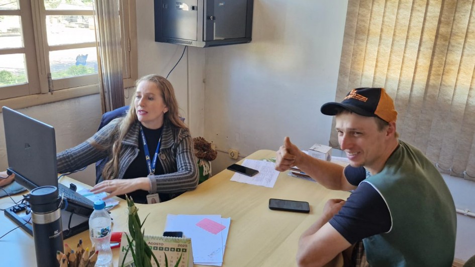 Uma foto dentro de um escritório municipal simples, em que uma mulher e um homem estão sentados em uma mesa de trabalho. A mulher, à esquerda, está sentada em frente a um computador, digitando, e parece estar ajudando o homem. Ela tem cabelos longos e loiros, e veste um casaco cinza escuro. Ela usa um crachá de identificação. O homem, à direita, está sorrindo para a câmera e fazendo um sinal de "joinha" com a mão direita. Ele usa um boné de beisebol (laranja e preto, com a marca "VZ") e um colete ou camisa regata verde sobre uma camisa preta.
A mesa de madeira clara contém vários objetos, incluindo um celular, papéis (incluindo uma folha rosa), uma garrafa térmica preta, garrafas de água mineral e um calendário de mesa que indica "AGOSTO 2020". O fundo mostra uma janela de madeira à esquerda, com persianas bege que deixam passar a luz do sol, e uma parede de cor clara. Acima e à esquerda, há um pequeno armário ou caixa preta fixada na parede.
