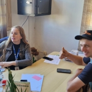 
Uma foto dentro de um escritório municipal simples, em que uma mulher e um homem estão sentados em uma mesa de trabalho. A mulher, à esquerda, está sentada em frente a um computador, digitando, e parece estar ajudando o homem. Ela tem cabelos longos e loiros, e veste um casaco cinza escuro. Ela usa um crachá de identificação. O homem, à direita, está sorrindo para a câmera e fazendo um sinal de "joinha" com a mão direita. Ele usa um boné de beisebol (laranja e preto, com a marca "VZ") e um colete ou camisa regata verde sobre uma camisa preta.

A mesa de madeira clara contém vários objetos, incluindo um celular, papéis (incluindo uma folha rosa), uma garrafa térmica preta, garrafas de água mineral e um calendário de mesa que indica "AGOSTO 2020". O fundo mostra uma janela de madeira à esquerda, com persianas bege que deixam passar a luz do sol, e uma parede de cor clara. Acima e à esquerda, há um pequeno armário ou caixa preta fixada na parede.
