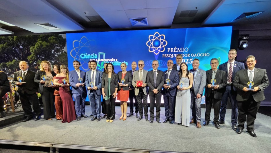 Grupo de 17 pessoas posa no palco segurando troféus do Prêmio Pesquisador Gaúcho 2025. Homens e mulheres estão alinhados diante de um grande painel azul com o tema “Ciência para reconstrução e resiliência”. A cena transmite um clima de celebração e reconhecimento à pesquisa científica.