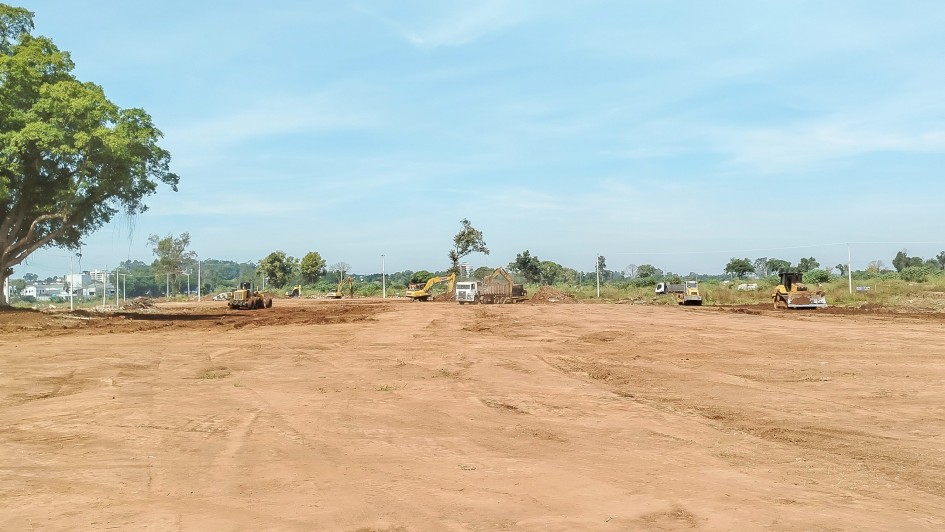 A foto mostra um terreno amplo de solo avermelhado. A área está limpa, sem vegetação, com marcas de máquinas no chão. Ao fundo, há alguns tratores e caminhões posicionados próximos a montes de terra.