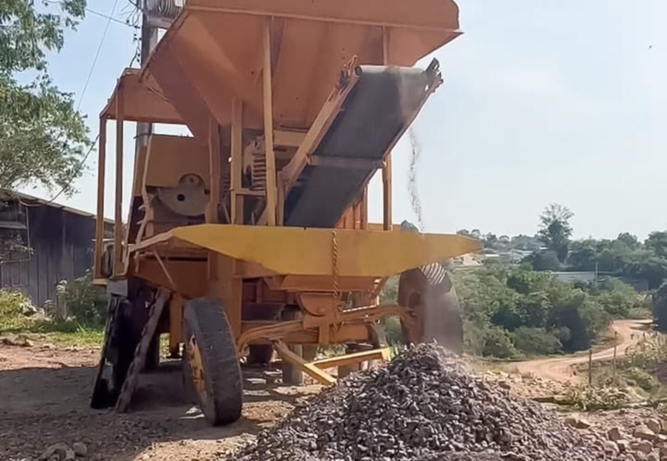 A foto mostra uma máquina de britagem de cor amarela posicionada sobre um terreno de terra. A estrutura possui uma correia transportadora inclinada, que despeja pedras britadas formando uma pilha no chão.