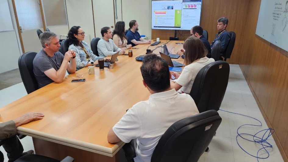 Foto de uma sala de reuniões, de paredes marrons e grande mesa retangular também marrom. Nove pessoas estão sentadas junto a essa mesa, e algumas olham para um slide projetado numa tela ao fundo.