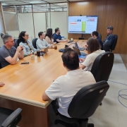 Foto de uma sala de reuniões, de paredes marrons e grande mesa retangular também marrom. Nove pessoas estão sentadas junto a essa mesa, e algumas olham para um slide projetado numa tela ao fundo.
