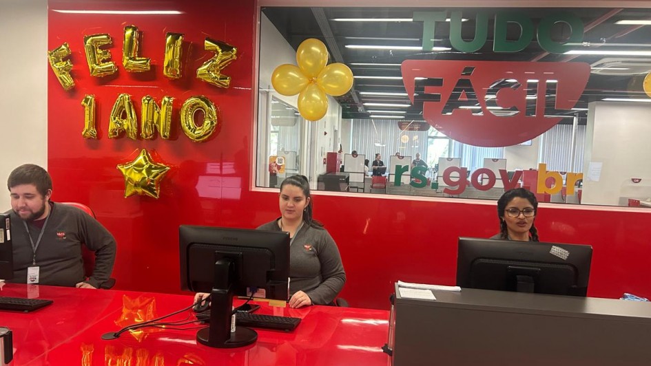 Imagem mostra, da esquerda para a direita, três pessoas no balcão de atendimentos do Tudo Fácil. Ao fundo, há decoração alusiva ao aniversário da agência