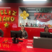 Imagem mostra, da esquerda para a direita, três pessoas no balcão de atendimentos do Tudo Fácil. Ao fundo, há decoração alusiva ao aniversário da agência
