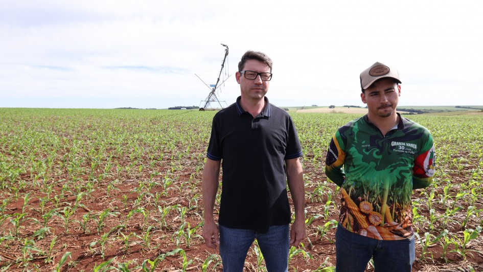 A foto mostra duas pessoas em pé sobre um campo agrícola com solo avermelhado e plantas jovens alinhadas em fileiras. Ao fundo, há um equipamento de irrigação do tipo pivô central, com estrutura metálica longa e aspersores pendurados.
