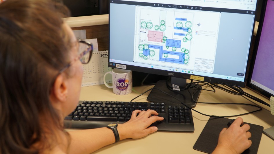 Imagem em plano fechado exibe uma servidora da Secretaria de Obras Públicas, de perfil, visualizando projeto de engenharia na tela em seu computador de trabalho.