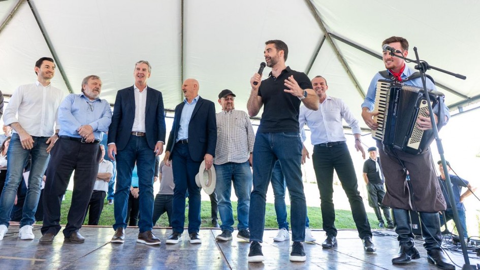A foto mostra um grupo de pessoas sobre um palco coberto por uma estrutura de lona branca. No centro, o governador fala ao microfone, enquanto outras pessoas estão alinhadas ao redor.