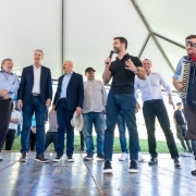 A foto mostra um grupo de pessoas sobre um palco coberto por uma estrutura de lona branca. No centro, o governador fala ao microfone, enquanto outras pessoas estão alinhadas ao redor.