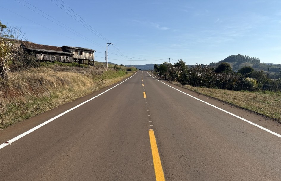 A imagem mostra uma estrada asfaltada de mão dupla em área rural, com faixa central amarela contínua e laterais brancas.