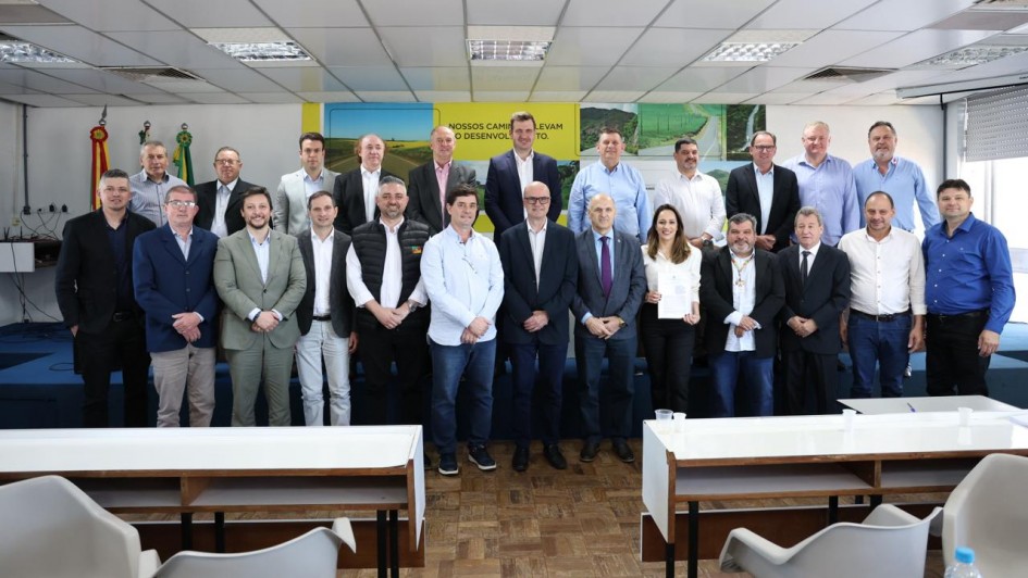 Grupo de cerca de 25 pessoas posa para foto em sala de reuniões iluminada, com bandeiras do Brasil e do Rio Grande do Sul ao fundo. No centro, autoridades estaduais e municipais seguram documentos de cooperação.