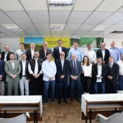 Grupo de cerca de 25 pessoas posa para foto em sala de reuniões iluminada, com bandeiras do Brasil e do Rio Grande do Sul ao fundo. No centro, autoridades estaduais e municipais seguram documentos de cooperação. 