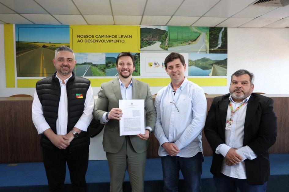 Quatro homens posam sorridentes diante de um painel amarelo com a frase “Nossos caminhos levam ao desenvolvimento”, ilustrado com imagens de rodovias e paisagens do Estado. O homem ao centro segura um documento oficial, simbolizando a assinatura dos termos de cooperação do Programa Horas-máquina.