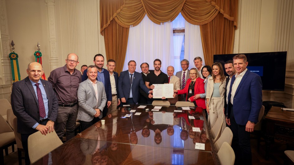 Uma foto em ambiente interno que captura um grupo de cerca de 16 pessoas (homens e mulheres) em pé, posando ao redor de uma grande e lustrosa mesa de reunião de madeira escura. O homem no centro da foto (de camisa preta) e a mulher ao lado dele (de blazer vermelho) estão segurando um documento estendido, possivelmente um termo de regulamentação ou lei, e todos estão olhando para a câmera. Os demais indivíduos vestem trajes sociais ou casuais de negócios, como ternos, blazers e camisas sociais.
O ambiente é formal e opulento, com paredes claras, cortinas de veludo marrom emoldurando uma janela ao fundo, um lustre no teto e objetos de decoração (incluindo faixas ou estandartes coloridos) no canto esquerdo. A sala está bem iluminada. O grupo parece ser formado por autoridades e membros do governo envolvidos na regulamentação de um programa de saúde.