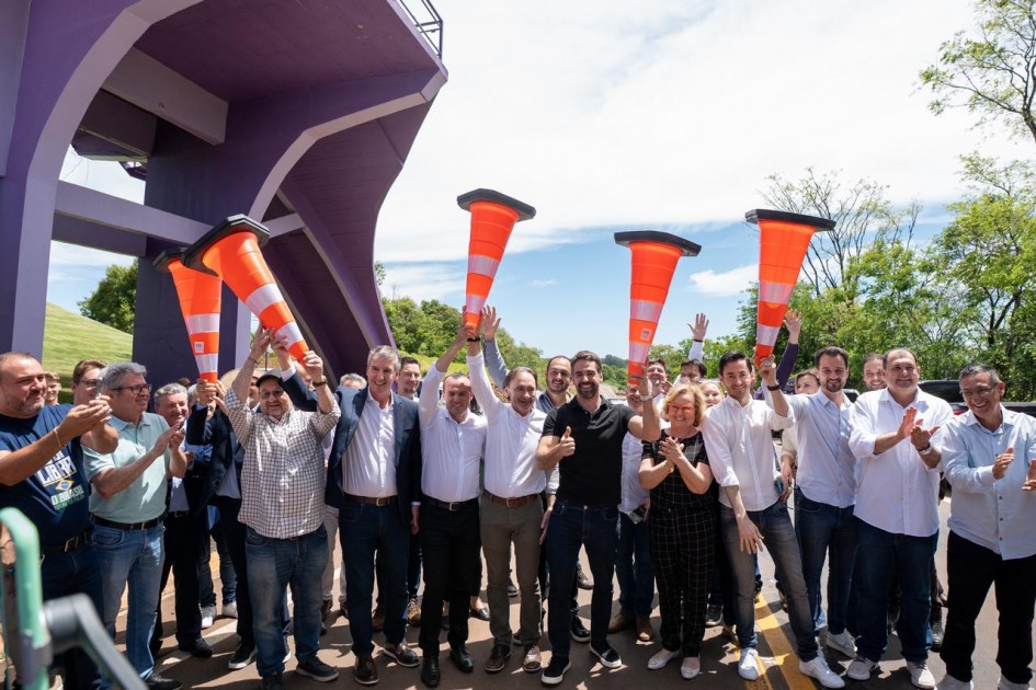 Uma foto externa, vibrante, de um grupo grande de pessoas (homens e algumas mulheres) celebrando um evento, possivelmente a inauguração ou o início de uma obra. O grupo está de pé, sorrindo e aplaudindo. Vários deles estão segurando cones de trânsito grandes, de cor laranja e branca, acima de suas cabeças como sinal de celebração.
No centro do grupo, um homem de camisa polo preta (o mesmo das fotos anteriores, Eduardo Leite) está fazendo um sinal de "joinha" com a mão direita, e a mulher ao lado dele está aplaudindo. A maioria dos homens veste camisas sociais ou camisetas.
O fundo é dominado pela estrutura de uma ponte ou viaduto, pintada de um tom roxo/lavanda escuro, que se eleva sobre a esquerda. O restante do fundo é composto por vegetação verde e densa e um céu claro com algumas nuvens. O chão é de asfalto ou terra batida.