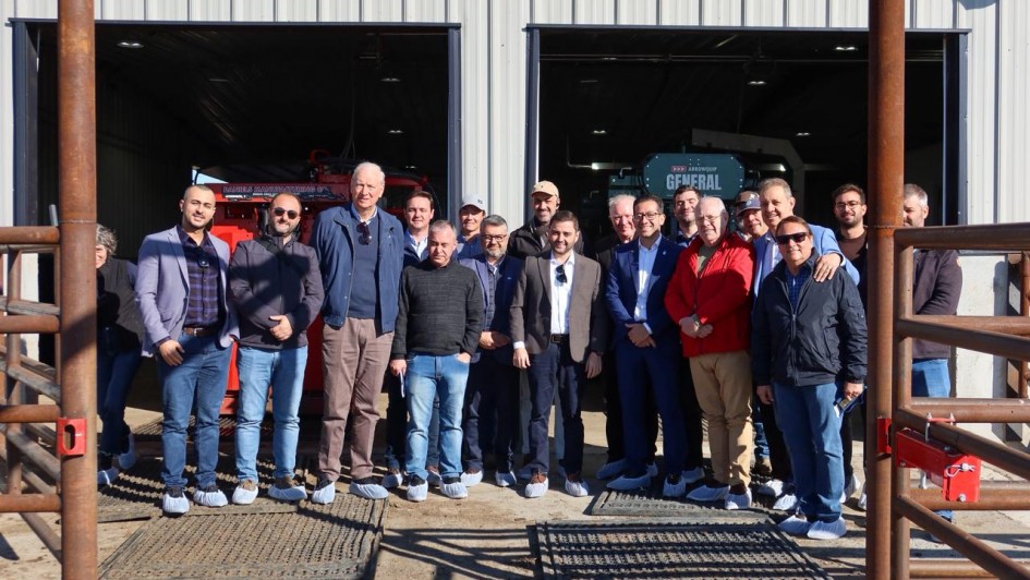 Uma foto externa de um grupo de cerca de 18 homens posando na frente da entrada aberta de um galpão. O grupo, vestindo roupas casuais de negócios ou casuais (muitos de jeans e blazers ou casacos), parece ser uma comitiva em visita. Todos usam protetores de sapatos (propés) de plástico azul-claro ou branco. Atrás deles, dentro do galpão, são visíveis grandes equipamentos agrícolas, incluindo uma máquina vermelha à esquerda e um equipamento verde escuro com a palavra "GENERAL" no centro. O galpão tem portas de correr de metal cinza. O grupo está em uma área de pátio com cercas de metal marrom de manejo de gado em primeiro plano, sugerindo que a visita é a uma instalação de produção animal ou agrícola de grande escala. O dia é de sol, com sombras projetadas no chão.