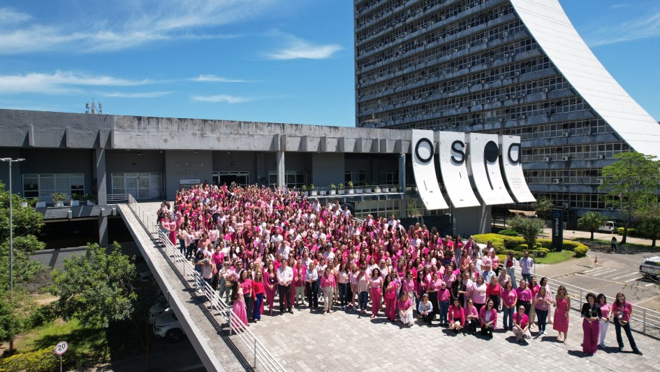 Uma foto aérea de um grande grupo de pessoas, predominantemente mulheres, vestidas em tons de rosa e branco, reunidas em uma esplanada ao ar livre em frente a um prédio. A foto foi tirada para marcar o "Outubro Rosa". O grupo está posicionado na rampa e na área pavimentada em frente a um edifício cinza de arquitetura moderna, com um letreiro que parece dizer "CAFF". Ao fundo e à direita, eleva-se um arranha-céu branco e curvo. O dia está ensolarado com céu azul. A legenda original da foto menciona "Servidores estaduais prestigiam foto coletiva do Outubro Rosa no CAFF" (Servidores públicos estaduais prestigiam foto coletiva do Outubro Rosa no CAFF).