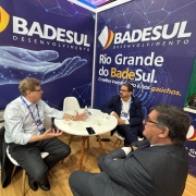 A imagem mostra três homens sentados ao redor de uma mesa redonda, conversando entre si. Ao fundo, há cartazes com a inscrição "Badesul Desenvolvimento" e "Rio Grande do Badesul". 