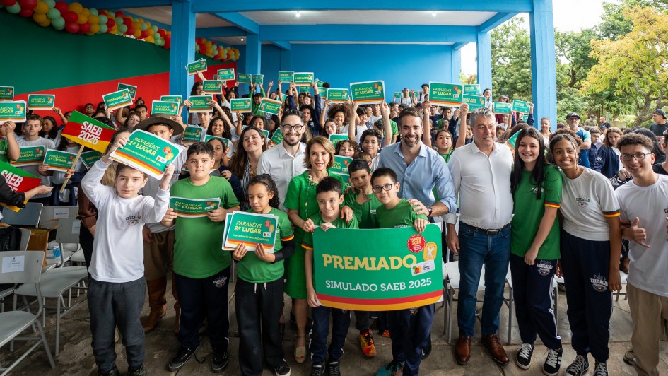 A imagem retrata um grupo numeroso de estudantes e adultos reunidos para uma foto coletiva, com o governador ao centro. Todos estão posicionados sob uma estrutura azul, segurando cartazes verdes com frases como “Premiado Simulado Saeb 2025”.