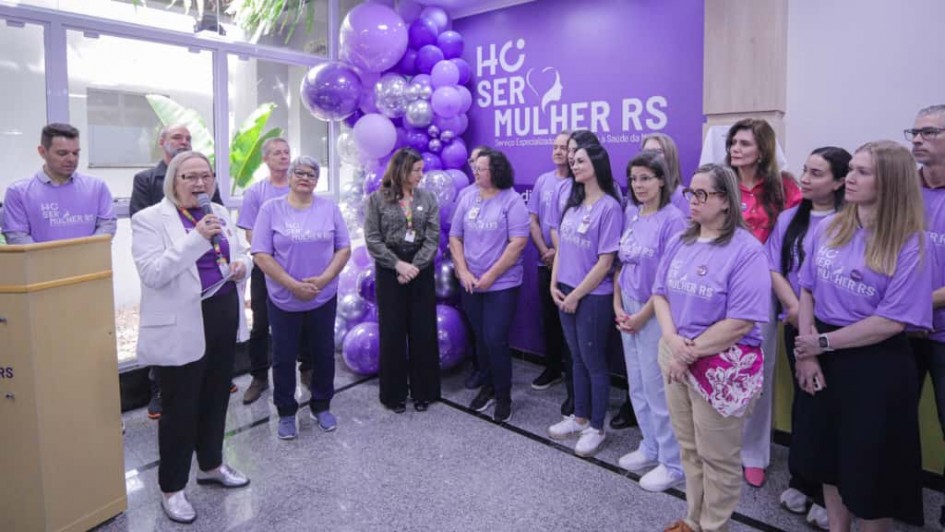 A imagem mostra a secretária falando ao microfone em pé, enquanto um grupo de mulheres acompanha a fala. Todas vestem roupas em tons de roxo com a marca do programa SERMulher RS. 