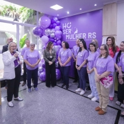 A imagem mostra a secretária falando ao microfone em pé, enquanto um grupo de mulheres acompanha a fala. Todas vestem roupas em tons de roxo com a marca do programa SERMulher RS. 