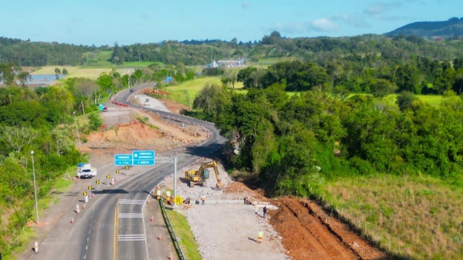 Imagem aérea mostra trecho da rodovia RSC-287 em obras, cercado por áreas de vegetação e campos verdes. O tráfego segue por uma das pistas, enquanto máquinas e trabalhadores atuam na construção de um novo trecho ao lado. Placas azuis indicam direções para “Candelária” e “Agudo”.