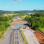 Imagem aérea mostra trecho da rodovia RSC-287 em obras, cercado por áreas de vegetação e campos verdes. O tráfego segue por uma das pistas, enquanto máquinas e trabalhadores atuam na construção de um novo trecho ao lado. Placas azuis indicam direções para “Candelária” e “Agudo”.