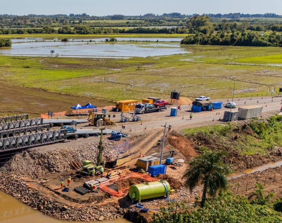 Imagem aérea mostra o canteiro de obras de uma ponte na RSC-287, com maquinário pesado, caminhões e contêineres de apoio. Há operários trabalhando e estruturas metálicas próximas ao leito de um rio. Ao fundo, aparecem campos alagados e áreas de vegetação.