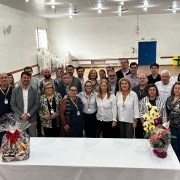 A imagem mostra um grupo de pessoas reunidas em pé, posando para a foto. Elas estão em uma sala grande, com uma mesa retangular com arranjos de flores na frente. 