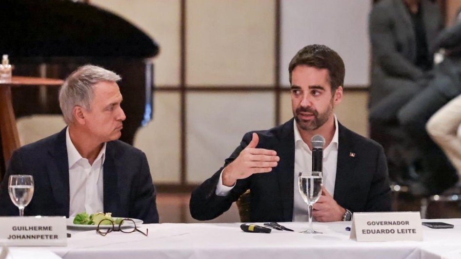 Dois homens sentados à mesa em reunião formal. Um deles fala e gesticula, o outro ouve com atenção. Ambos usam traje social, com nome em placas à frente. Ambiente elegante e profissional.