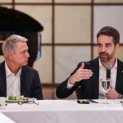 Dois homens sentados à mesa em reunião formal. Um deles fala e gesticula, o outro ouve com atenção. Ambos usam traje social, com nome em placas à frente. Ambiente elegante e profissional.