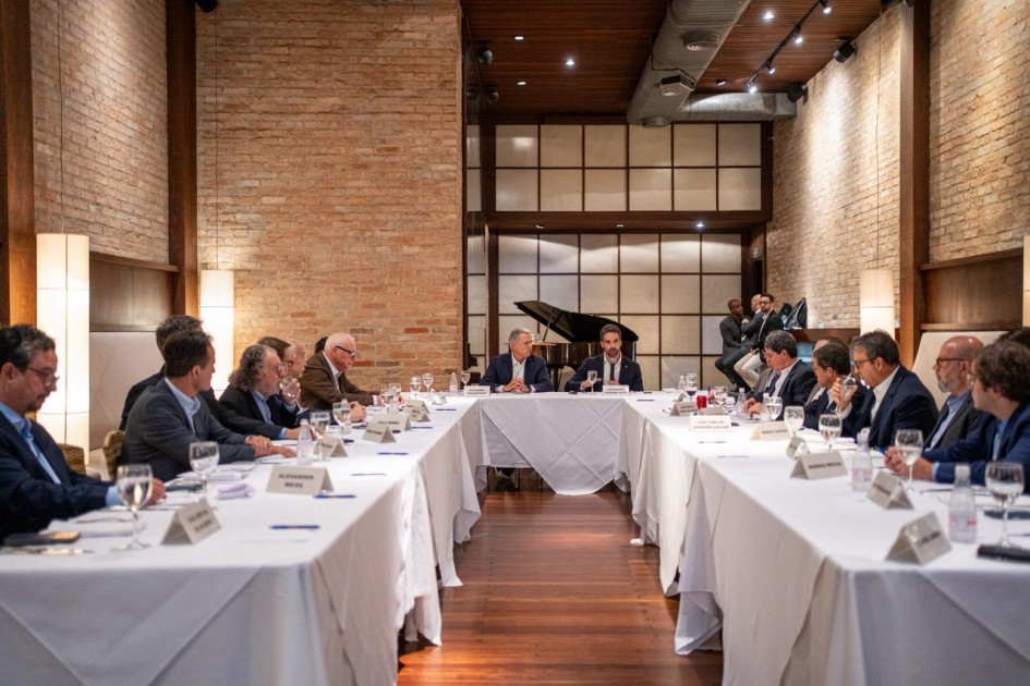 Reunião formal em sala elegante com mesa em formato de U. Pessoas de terno discutem assuntos profissionais, com placas de nome e materiais à frente. Ao fundo, há piano e bateria.