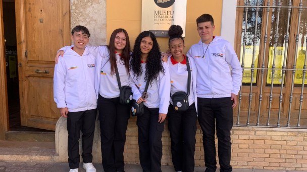 Cinco jovens posam lado a lado, sorrindo, diante da entrada de um edifício identificado como “Museu Comarcal d’Horta Sud”. Eles vestem calças pretas e agasalhos brancos com o logotipo “Partiu Futuro Reconstrução”. Três deles carregam pequenas bolsas transversais pretas. Atrás, há uma porta de madeira e uma janela com grades metálicas.