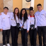 Cinco jovens posam lado a lado, sorrindo, diante da entrada de um edifício identificado como “Museu Comarcal d’Horta Sud”. Eles vestem calças pretas e agasalhos brancos com o logotipo “Partiu Futuro Reconstrução”. Três deles carregam pequenas bolsas transversais pretas. Atrás, há uma porta de madeira e uma janela com grades metálicas.