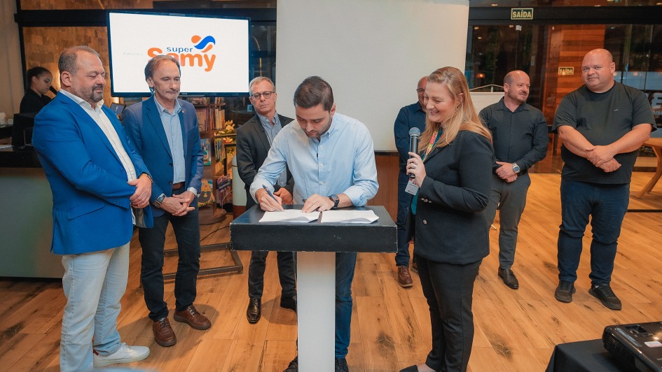 O vice-governador Gabriel Souza está assinando um documento sobre um púlpito, enquanto uma mulher ao seu lado segura um microfone. Outras seis pessoas, em pé, observam a ação.