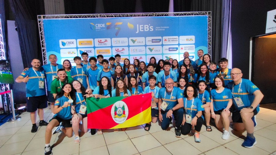 Foto mostra grupo de 60 pessoas, entre estudantes e representantes da comissão técnica, posando com medalhas e uma bandeira do Estado, tendo ao fundo o banner oficial dos Jogos Escolares Brasileiros