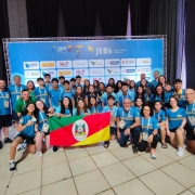Foto mostra grupo de 60 pessoas, entre estudantes e representantes da comissão técnica, posando com medalhas e uma bandeira do Estado, tendo ao fundo o banner oficial dos Jogos Escolares Brasileiros