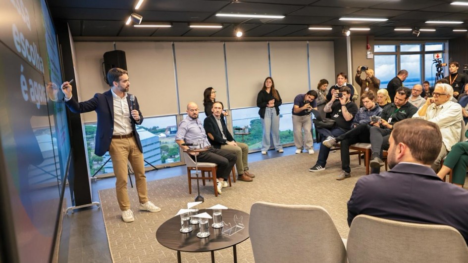 Uma foto em ambiente interno que captura uma apresentação ou lançamento de projeto em uma sala moderna. No canto esquerdo, de pé, um homem (o mesmo visto em outras fotos, Eduardo Leite) veste blazer escuro sobre camisa branca e calças claras, e está gesticulando com a mão direita em direção a uma grande tela escura que exibe texto em cor vibrante e letras grandes. No restante da sala, em ângulo diagonal, uma audiência de cerca de 15 a 20 pessoas está sentada em cadeiras ou poltronas, ou de pé na parte de trás, observando a apresentação. O público é composto por homens e mulheres, alguns de terno e outros com roupas casuais. No centro, há uma pequena mesa redonda preta com copos d'água e microfones de lapela ou crachás. O fundo apresenta uma grande janela horizontal que mostra uma paisagem urbana. O teto é preto com iluminação de trilhos. A foto sugere o lançamento da concessão do serviço de bilhetagem eletrônica do transporte metropolitano.