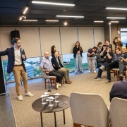 Uma foto em ambiente interno que captura uma apresentação ou lançamento de projeto em uma sala moderna. No canto esquerdo, de pé, um homem (o mesmo visto em outras fotos, Eduardo Leite) veste blazer escuro sobre camisa branca e calças claras, e está gesticulando com a mão direita em direção a uma grande tela escura que exibe texto em cor vibrante e letras grandes. No restante da sala, em ângulo diagonal, uma audiência de cerca de 15 a 20 pessoas está sentada em cadeiras ou poltronas, ou de pé na parte de trás, observando a apresentação. O público é composto por homens e mulheres, alguns de terno e outros com roupas casuais. No centro, há uma pequena mesa redonda preta com copos d'água e microfones de lapela ou crachás. O fundo apresenta uma grande janela horizontal que mostra uma paisagem urbana. O teto é preto com iluminação de trilhos. A foto sugere o lançamento da concessão do serviço de bilhetagem eletrônica do transporte metropolitano.