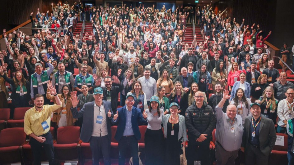 A imagem mostra um auditório lotado. Na primeira fileira, em pé, há oito pessoas sorrindo e acenando com as mãos. Ao fundo, a plateia está com os braços erguidos, posando para a foto.