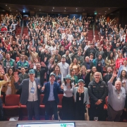 A imagem mostra um auditório lotado. Na primeira fileira, em pé, há oito pessoas sorrindo e acenando com as mãos. Ao fundo, a plateia está com os braços erguidos, posando para a foto. 