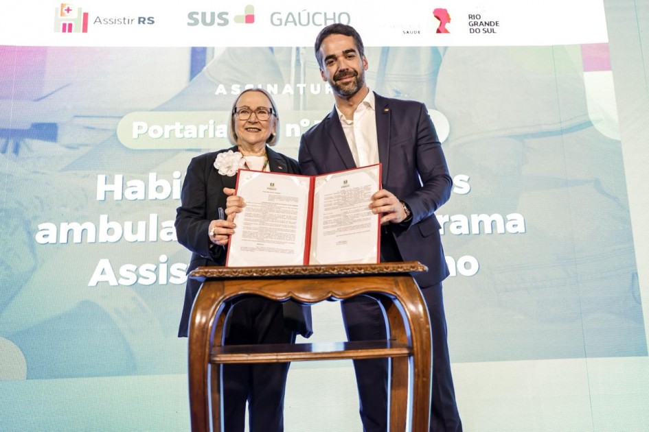 Dois representantes em evento oficial mostram documento assinado sobre saúde pública. Estão em frente a tela com logotipos e texto do programa Assistir RS, ligado ao SUS e ao governo do Rio Grande do Sul. Ambiente formal e institucional.