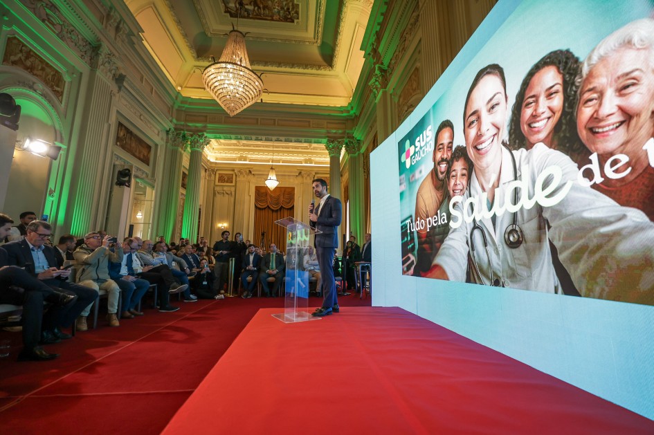 Homem discursa no palco com tapete vermelho, no salão elegante. Atrás dele, tela mostra imagem de profissionais de saúde e idosos sorrindo, com texto sobre o SUS e saúde pública. O público está sentado e atento.