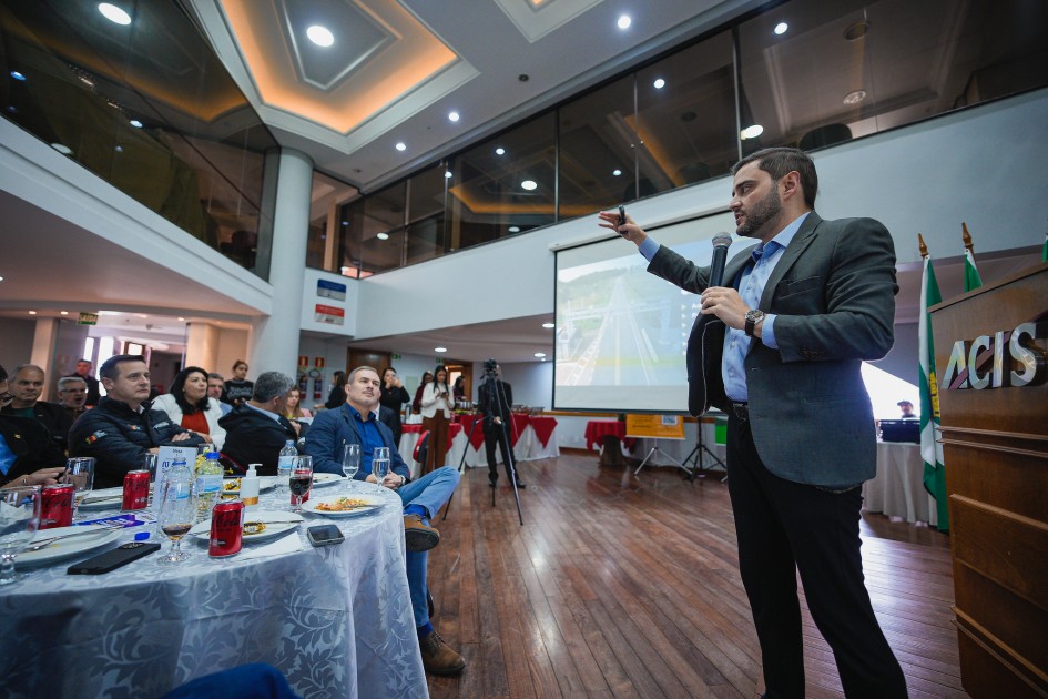 O vice-governador Gabriel Souza aparece de perfil, segurando um microfone e apontando para uma tela de projeção. À frente dele, há mesas com garrafas de água, copos e latas de refrigerante, onde estão sentadas várias pessoas assistindo à apresentação. Ao fundo, há bandeiras e um projetor ligado.